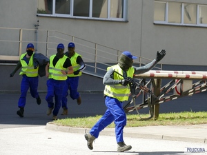 4 zamaskowanych mężczyzn w odblaskowych kamizelkach z karabinami w ręku biegną przed siebie, jeden z nich coś pokazuje za kadrem lewą ręką