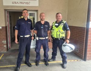 Na zdjęciu trzech uśmiechniętych policjantów stojących przed budynkiem szpitala