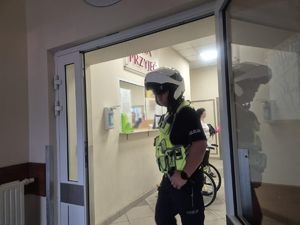 Na zdjęciu policjant w białym kasku na głowie, stojący w szpitalu przed izbą przyjęć