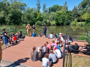 Policjant i strażacy prowadzą spotkanie z młodzież na pomoście przy rzece.