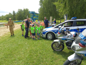 Dwóch policjantów i strażak prezentują grupie dzieci radiowóz i motocykle policyjne.