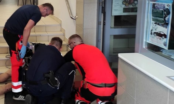 policjant i dwaj ratownicy medyczni przy mężczyźnie leżącym przy schodach na korytarzu w budynku
