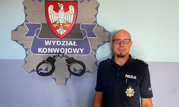 Policjant mł. asp. Szymon Karolewicz w mundurze na tle logo Wydziału Konwojowego KWP w Poznaniu