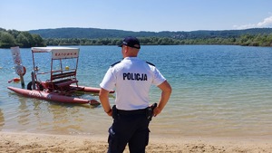 Policjant stoi na plaży odwrócony w kierunku zbiornika wodnego, który jest w tle.