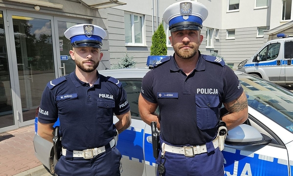dwóch umundurowanych policjantów stoi przy radiowozie