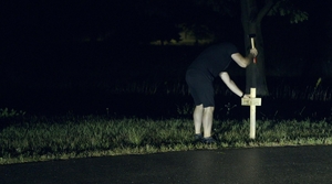 kadr z filmu: mężczyzna wbija młotkiem krzyż przy drodze