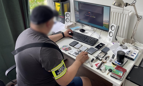 funkcjonariusz cbzc siedzi przy stanowisku komputerowym, na stoliku leżą różne drobne przedmioty zabezpieczone przez policjantów: nośniki pamięci, zegarek, dowody tożsamości