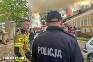 na zdjęciu policjant rozmawia ze strażakiem, w tle trwa akcja gaśnicza budynku