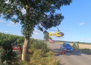 miejsce wypadku: na skraju pola kukurydzy stoi motocykl, na drodze obok stoi radiowóz policyjny, a w głębi wóz strażacki. Nad drogą wisi śmigłowiec pogotowia ratunkowego