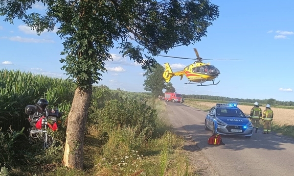 miejsce wypadku: na skraju pola kukurydzy stoi motocykl, na drodze obok stoi radiowóz policyjny, a w głębi wóz strażacki. Nad drogą wisi śmigłowiec pogotowia ratunkowego