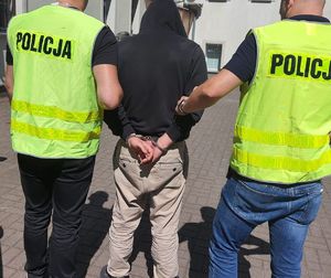 dwaj policjanci w żółtych kamizelkach z napisem Policja na plecach prowadzą zatrzymanego mężczyznę