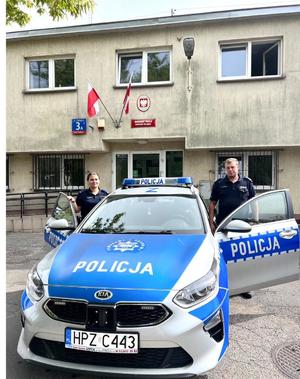 policjant i policjantka stoją przy radiowozem, w tle budynek komisariatu policji