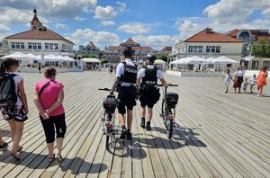 dwaj policjanci z patrolu rowerowego prowadzą rowery idąc po molo w Sopocie, obok nich i w tle turyści