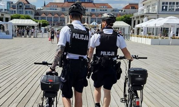 dwaj policjanci z patrolu rowerowego prowadzą rowery idąc po molo w Sopocie, w tle turyści