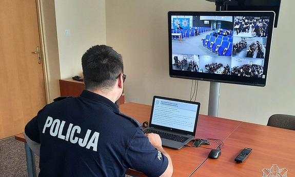 policjant siedzi przy stoliku, na którym stoi laptop, przed nim na stojaku wisi ekran