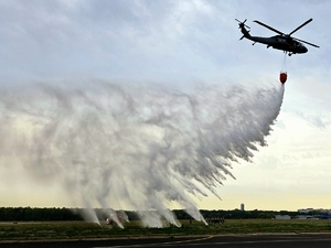 śmigłowiec w locie z podwieszonym zbiornikiem Bambi Bucket, z którego wylewa się woda