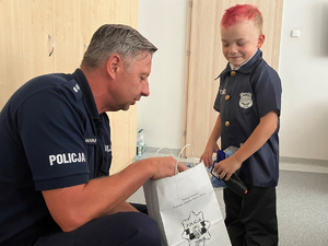 policjant kuca przy małym chłopcu. Z papierowej torby policjant wyciąga upominek.