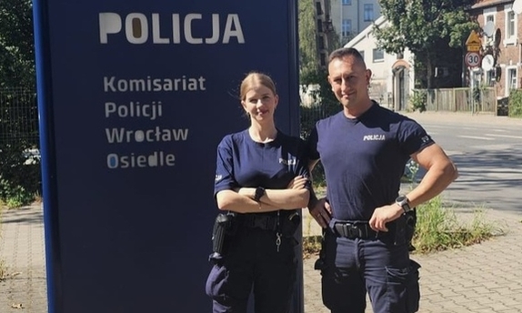 dwoje umundurowanych policjantów stojących przy tablicy informacyjnej z napisem Komisariat Policji Wrocław Osiedle