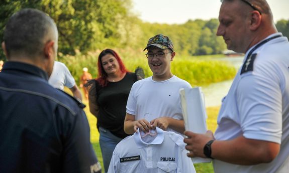 Chłopak uśmiecha się do aparatu, w ręku ma bluzkę ze swoim nazwiskiem i napisem policja