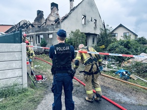 na zdjęciu umundurowany policjant i strażacy pracujący na miejscu zdarzenia, w tle spalony dom