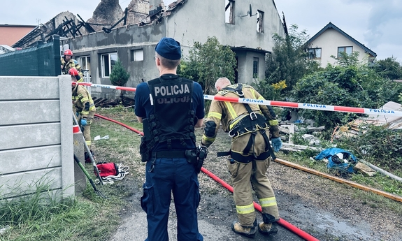 na zdjęciu umundurowany policjant i strażacy pracujący na miejscu zdarzenia, w tle spalony dom