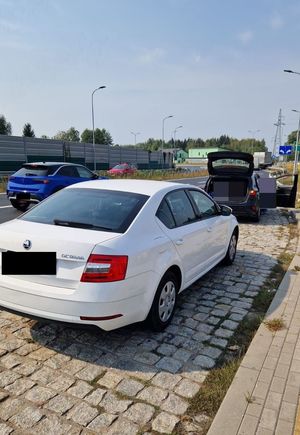 zdjęcie przedstawia samochód osobowy koloru białego marki Skoda a w tle są widoczne inne pojazdy. Auto stoi na parkingu