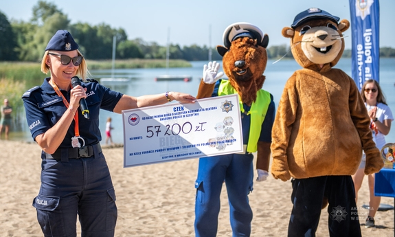 Komendant-Rektor Akademii Policji w Szczytnie trzyma symboliczny czek ze wpisaną kwotą 57200 złotych, obok niej stoją dwie maskotki policyjne. W tle plaża nad jeziorem.