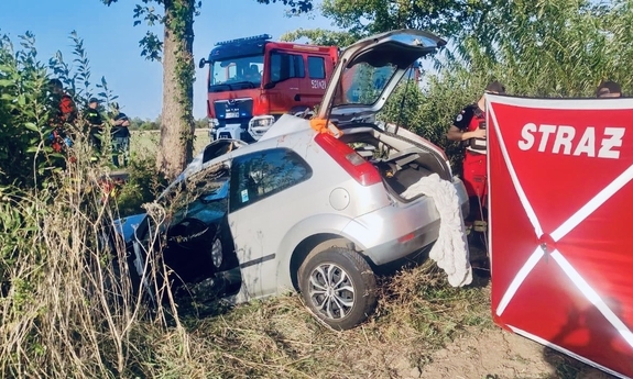 miejsce wypadku drogowego, na zdjęciu samochód w rowie. W tle wóz strażacki, strażacy i policjanci