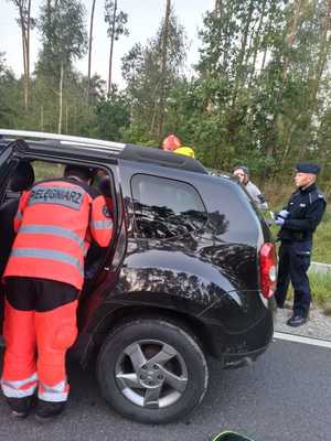 ratownik medyczny zagląda do czarnego samochodu przez otwarte tylne drzwi, obok stoi policjant i inne osoby