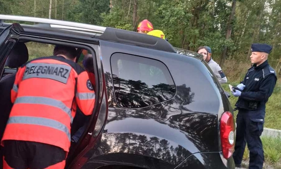 ratownik medyczny zagląda do czarnego samochodu przez otwarte tylne drzwi, obok stoi policjant i inne osoby