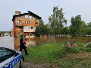 policjant niesie worek z piaskiem, w tle podtopiony budynek