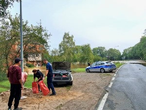 policjant pomaga napełniać worki z piaskiem, w tle radiowóz zaparkowany na poboczu