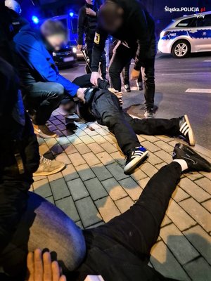 mężczyzna leżący na chodniku przytrzymywany przez nieumundurowanych policjantów. Obok nogi drugiego z zatrzymanych mężczyzn. W tle radiowozy policyjne