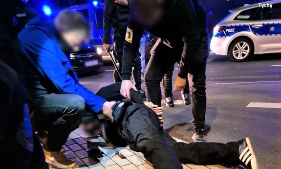 mężczyzna leżący na chodniku przytrzymywany przez nieumundurowanych policjantów. Obok nogi drugiego z zatrzymanych mężczyzn. W tle radiowozy policyjne