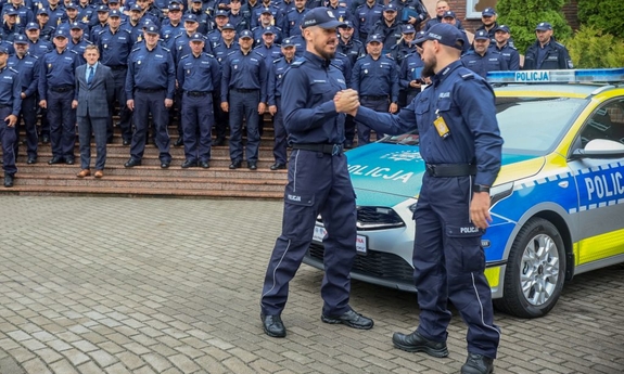 Na pierwszym planie dwaj policjanci podają sobie ręce stojąc obok radiowozu. W tle grupa policjantów na schodach