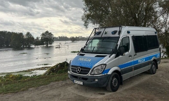 Radiowóz policyjny typu bus stoi na brzegu zbiornika wodnego.