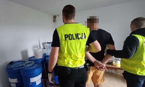 Dwaj policjanci z zatrzymanym mężczyzną w pomieszczeniu, pod ścianą stoją niebieskie beczki.