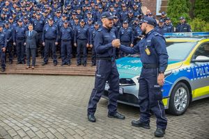 policjanci stoją przed radiowozem i przybijają sobie piątkę.