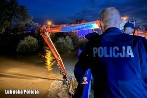 noc, policjant stoi na moście i patrzy na koparkę, która coś kopie.