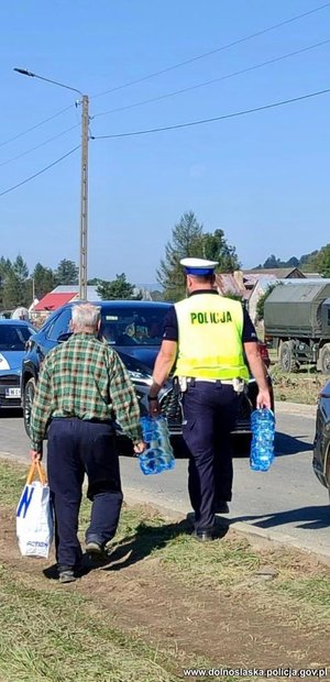 policjant niesie wodę, za nim idzie starszy mężczyzna, który też niesie wodę