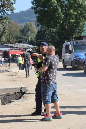 mężczyzna pyta o coś policjanta, w tle zniszczone miasto i wóz strażacki.