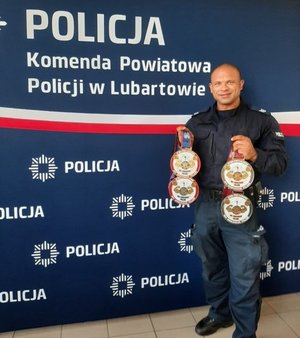 Na zdjęciu umundurowany policjant stoi na tle granatowej ścianki z napisem Policja, Komenda Powiatowa Policji w Lubartowie. Policjant w ręku trzyma zdobyte medale.