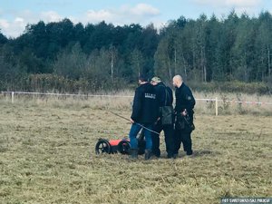 Policjanci w trakcie poszukiwań na trawiastym terenie, w tle las.