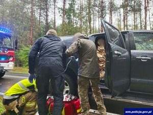 Miejsce zdarzenia drogowego. Trzej mężczyźni przy samochodzie udzielają pomocy poszkodowanemu.