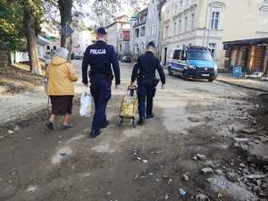 Dwaj umundurowani policjanci pomagają nieść seniorce produkty żywnościowe.