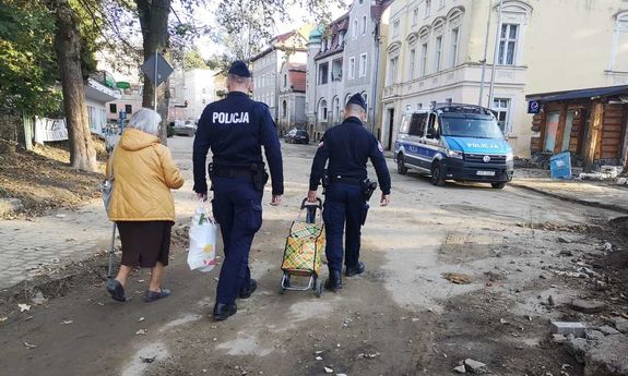 Dwaj umundurowani policjanci pomagają nieść seniorce produkty żywnościowe.