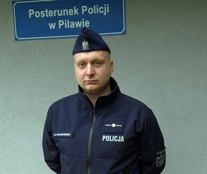 policjant umundurowany stoi na tle tablicy Posterunek Policji w Pilawie.