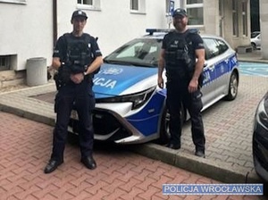 Dwaj policjanci stoją przed radiowozem.