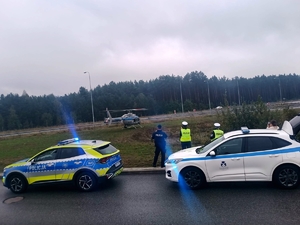 Na pierwszym planie radiowóz policyjni i biały samochód typu suv, obok którego stoją policjanci. W głębi śmigłowiec stoi na trawiastym terenie.