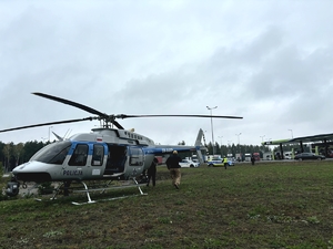 Śmigłowiec stoi na trawiastym terenie, w głębi policjant przy radiowozie oraz stacja benzynowa.